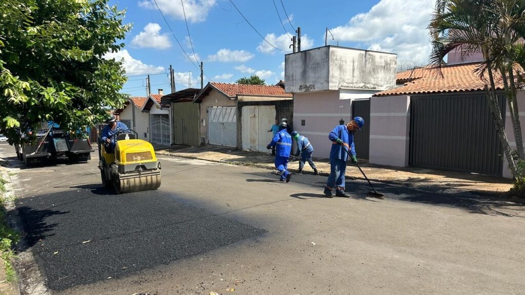 A imagem mostra uma cena de trabalho na manutenção de uma rua em um dia ensolarado. Vários trabalhadores estão vestidos com uniformes azuis e capacetes, realizando a pavimentação asfáltica. Um dos trabalhadores está operando um rolo compactador amarelo, que está passando sobre uma nova camada de asfalto escuro recém-aplicado na rua. Outros dois trabalhadores estão usando vassouras para espalhar e alisar o asfalto quente. Ao fundo, há um caminhão, provavelmente utilizado para transportar o material asfáltico. A rua parece estar em uma área residencial, com casas alinhadas de um lado, todas com telhados de cerâmica e portões fechados. O céu está azul com algumas nuvens brancas, e a luz do sol forte cria sombras visíveis no chão. A cena transmite uma sensação de atividade e trabalho em progresso, com cores que contrastam entre o asfalto escuro, o amarelo do equipamento, o azul dos uniformes e os tons terrosos das casas.