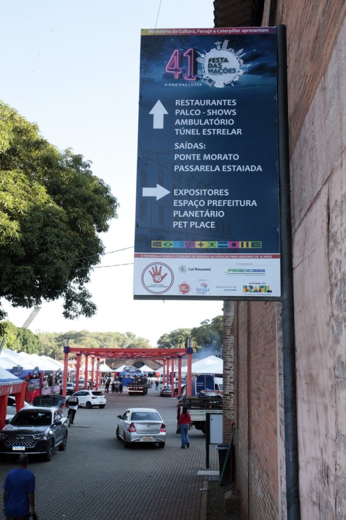 A imagem apresenta a sinalização informativa da "41ª Festa das Nações", evento realizado em Piracicaba, fixada em uma parede de tijolos. A placa detalha a localização de diversas atrações, como restaurantes, palcos, o "Túnel Estelar" e até um "Pet Place", além de indicar as saídas para a Ponte Morato e a Passarela Estaiada. O cenário ao fundo mostra um dia ensolarado, com uma estrutura metálica decorativa, tendas brancas montadas e o movimento de pedestres e veículos, característico de um evento público ao ar livre