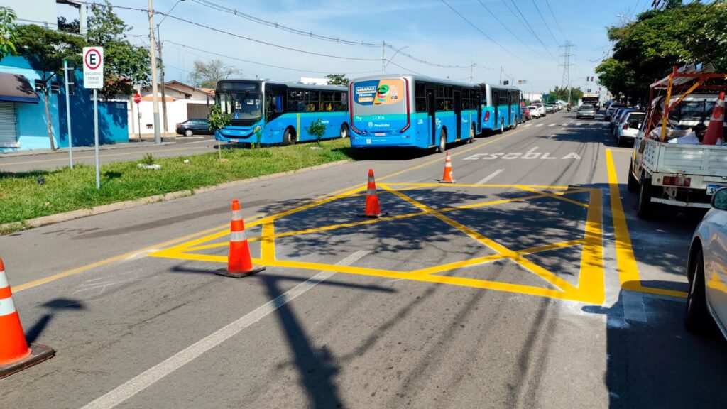 A imagem mostra uma cena urbana em uma rua com ônibus estacionados à esquerda. No chão, há uma pintura amarela formando um retângulo com linhas diagonais, indicando uma área de restrição de estacionamento, provavelmente para garantir a segurança próxima a uma escola, já que está escrito "ESCOLA" no asfalto. Em cima dessa área estão posicionados cones de trânsito laranja para delimitar o espaço. Dois ônibus azuis estão parados, um deles com um anúncio escrito "OBRAMAX" e o número "12306". À direita, há um veículo branco estacionado. Ao fundo, é possível ver um prédio azul e uma placa de estacionamento exclusivo para ônibus urbanos. A cena ocorre em um dia ensolarado, com sombras visíveis dos cones e dos fios elétricos na rua.