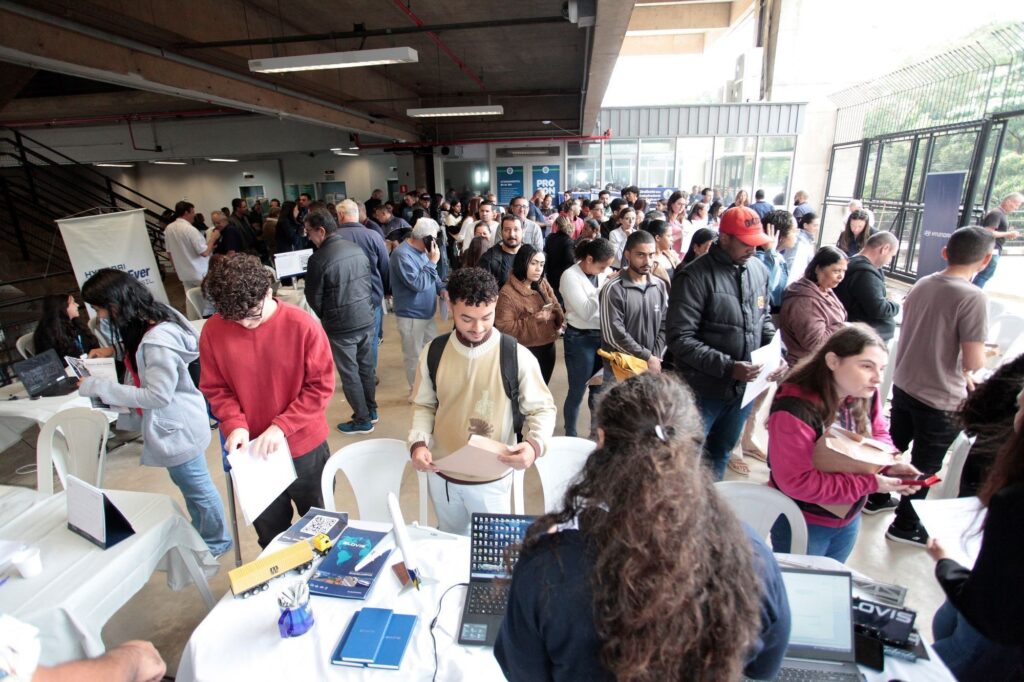 A imagem mostra um evento movimentado, provavelmente uma feira de empregos ou recrutamento, realizado em um espaço interno amplo com estrutura industrial, como teto alto e vigas expostas. Muitas pessoas estão presentes, a maioria delas parecem ser jovens candidatos, algumas em filas, outras conversando ou consultando materiais. Em primeiro plano, há uma mesa de atendimento com um tecido branco, onde estão dois laptops, um pequeno modelo de caminhão, folhetos promocionais da marca "SLOVIS" e alguns cadernos. Uma mulher de cabelos longos e encaracolados está de costas, aparentemente ajudando os visitantes. Ao fundo, a multidão se estende pelo local, com pessoas de diversas idades e etnias. No ambiente, podem ser vistos banners de empresas como Hyundai e PROCON, indicando que essas organizações provavelmente estão participando do evento. A iluminação é uma combinação de luz natural e artificial, criando um clima profissional e dinâmico típico de feiras de emprego.