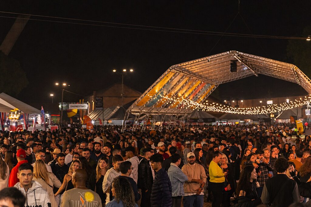 Esta imagem retrata uma cena noturna movimentada em um festival ou grande evento ao ar livre, capturando a energia de uma multidão concentrada. Aqui estão os principais detalhes observados: Ambiente e Iluminação: O cenário é dominado por uma grande estrutura metálica de palco (treliça) suspensa, decorada com fileiras de luzes amarelas, que criam uma atmosfera festiva e iluminam a área. O contraste entre o céu escuro da noite e as luzes quentes confere ao local um aspecto aconchegante, apesar da aglomeração. Multidão: O espaço está lotado de pessoas em pé, conversando ou observando o movimento, típico de shows ou festivais. Em primeiro plano, destacam-se vários indivíduos, incluindo um homem jovem de casaco com capuz no canto inferior esquerdo que parece pensativo. Contexto: Ao fundo, é possível notar tendas brancas e sinalizações (como uma placa indicando "EMPRÉSTIMO"), sugerindo a presença de uma área comercial ou de serviços dentro do evento. A foto transmite a sensação de estar imerso no local, com um clima de "caos organizado" e descontração característico desse tipo de celebração pública.