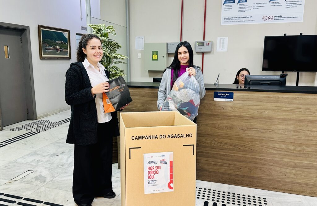 A imagem mostra duas jovens em um saguão, provavelmente na recepção da Câmara Municipal de Piracicaba, participando de uma Campanha do Agasalho. Elas estão sorrindo, segurando bolsas com roupas para doação e posicionadas ao lado de uma caixa de papelão preparada para receber as contribuições, com cartazes incentivando a solidariedade. O ambiente é típico de um prédio público, com balcão de atendimento ao fundo e sinalizações informativas nas paredes. É uma cena bem positiva de conscientização e apoio ao próximo