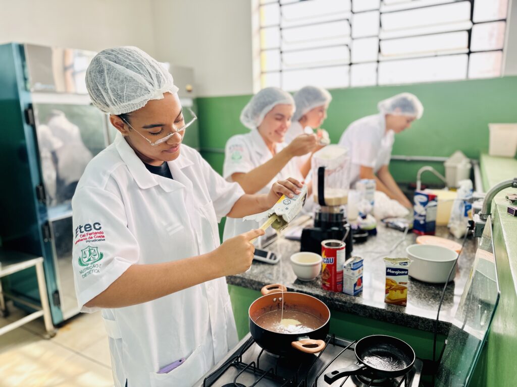 A imagem mostra um ambiente de cozinha profissional, provavelmente uma cozinha experimental de uma instituição de ensino, onde quatro alunas estão vestidas com jalecos brancos e toucas para higiene. A aluna em destaque, em primeiro plano, está concentrada enquanto despeja um líquido de uma embalagem em um recipiente de metal (panela ou tigela) sobre um fogão. Ela usa óculos e está vestindo o jaleco com o emblema da instituição "Etec Cel. Fernando Febeliano da Costa Piracicaba" e a área de estudo "Nutrição e Dietética". Ao fundo, outras três alunas também estão envolvidas em atividades culinárias, como mexer ou preparar ingredientes, todas igualmente protegidas com jalecos e toucas. O ambiente é bem iluminado, com paredes verdes e bancadas de granito, além de equipamentos e utensílios típicos de cozinha profissional, como liquidificadores e panelas. Na bancada, há uma embalagem da marca "Marajoara", que parece ser um ingrediente usado na preparação. A cena transmite um ambiente de aprendizado prático, com foco em nutrição e dietética, onde as estudantes aplicam conhecimentos em um laboratório de alimentos.