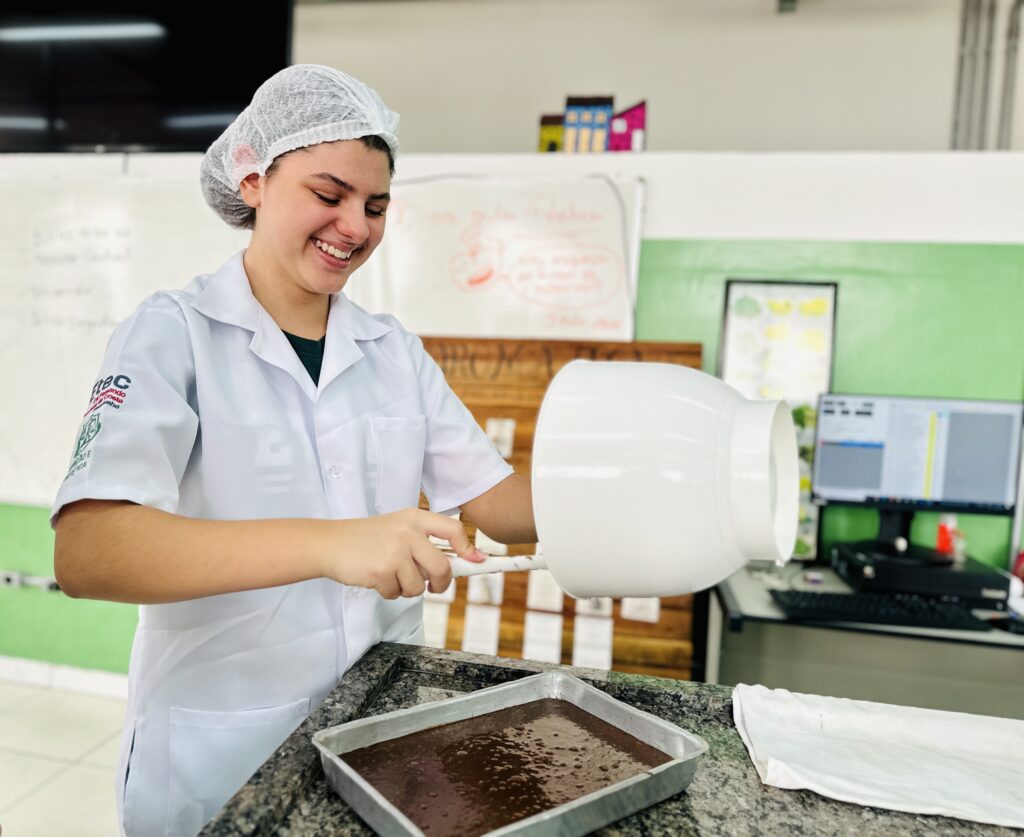 A imagem mostra uma jovem mulher em um ambiente que parece ser uma cozinha profissional ou laboratório de alimentos. Ela está sorrindo enquanto despeja uma massa de chocolate espessa e escura de uma tigela branca grande para uma forma retangular de metal. A mulher veste um jaleco branco e uma touca descartável, indicando que está em um ambiente de trabalho ou treinamento que exige higiene, como uma cozinha industrial ou laboratório. Ao fundo, há um quadro branco com anotações escritas, um monitor de computador sobre uma mesa e uma parede verde. O ambiente está bem iluminado, com luz clara destacando a textura da massa de chocolate e os detalhes do local. No jaleco, há o logo "Ftec", que provavelmente indica a instituição ou empresa relacionada ao local. A expressão da mulher é alegre e demonstra que ela está satisfeita com o que está fazendo.