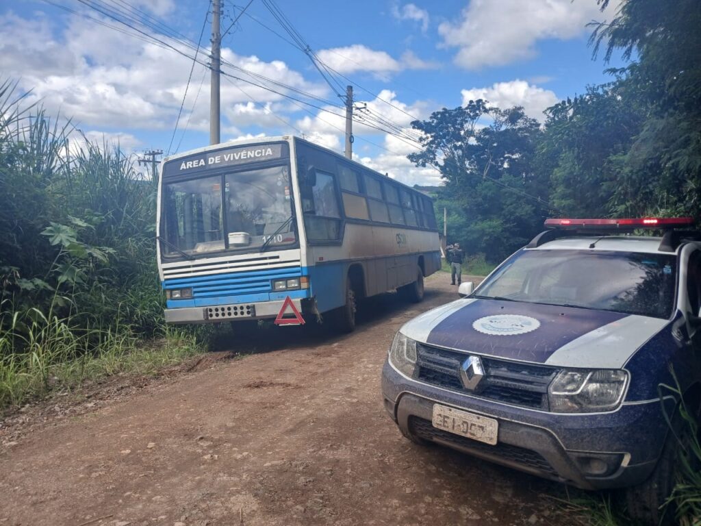 A imagem mostra uma estrada de terra em uma área rural ou semi-rural, cercada por vegetação densa e árvores ao fundo. À esquerda da imagem, está estacionado um ônibus antigo, branco com detalhes em azul, com a inscrição "ÁREA DE VIVÊNCIA" na parte frontal, indicando que provavelmente o veículo foi adaptado para servir como espaço de convivência. O número "140" está visível na parte frontal do ônibus. À direita, mais próximo do primeiro plano, está um carro da polícia, um SUV com pintura azul e branca, com sirenes ligadas (luzes vermelha e azul piscando). Um policial está parado próximo ao ônibus e ao carro, aparentemente verificando ou atendendo a situação. O céu está parcialmente nublado, com uma mistura de nuvens brancas e céu azul. A cena sugere uma intervenção policial ou algum tipo de fiscalização em uma área remota. A estrada de terra e o ambiente ao redor indicam que o local é afastado e cercado pela natureza.