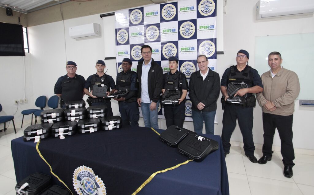 A imagem mostra um grupo de oito pessoas, entre homens e mulheres, posando para uma foto oficial em um ambiente interno. Eles estão alinhados atrás de uma mesa coberta com um pano azul escuro, sobre a qual estão dispostos vários estojos pretos de armas de fogo, alguns abertos, mostrando pistolas dentro. Ao fundo, há um painel com a logo da Prefeitura de Piracicaba e da Guarda Civil Metropolitana, indicando que o evento está relacionado a essa instituição. Algumas pessoas vestem uniformes táticos da Guarda Civil Metropolitana, enquanto outras estão com roupas civis. Todos olham para a câmera, e alguns seguram os estojos abertos, exibindo as armas. A iluminação é clara e uniforme, típica de um ambiente de escritório ou sala de reuniões. A cena aparenta ser uma apresentação formal de novas armas para a Guarda Civil Metropolitana de Piracicaba.
