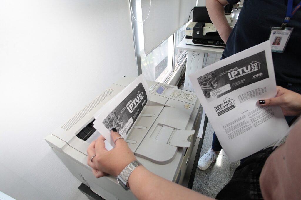 A imagem mostra um ambiente de escritório onde duas pessoas estão manuseando documentos impressos relacionados ao IPTU. * Em primeiro plano, uma pessoa segura uma folha de papel impressa que acaba de sair de uma impressora a laser de mesa. O documento destaca o título NOVO IPTU 2026 e o logotipo da prefeitura de Piracicaba. * A outra pessoa, à direita, segura um documento idêntico, exibindo também o texto NOVA PGV. NOVO IPTU. Ela utiliza um crachá de identificação com o nome "LAURA". * Ao fundo, é possível ver parte de uma mesa com outra impressora (HP) e uma janela com persianas brancas, indicando um local de trabalho administrativo. O foco principal é a atividade de organização e conferência dos boletos de pagamento do imposto municipal.