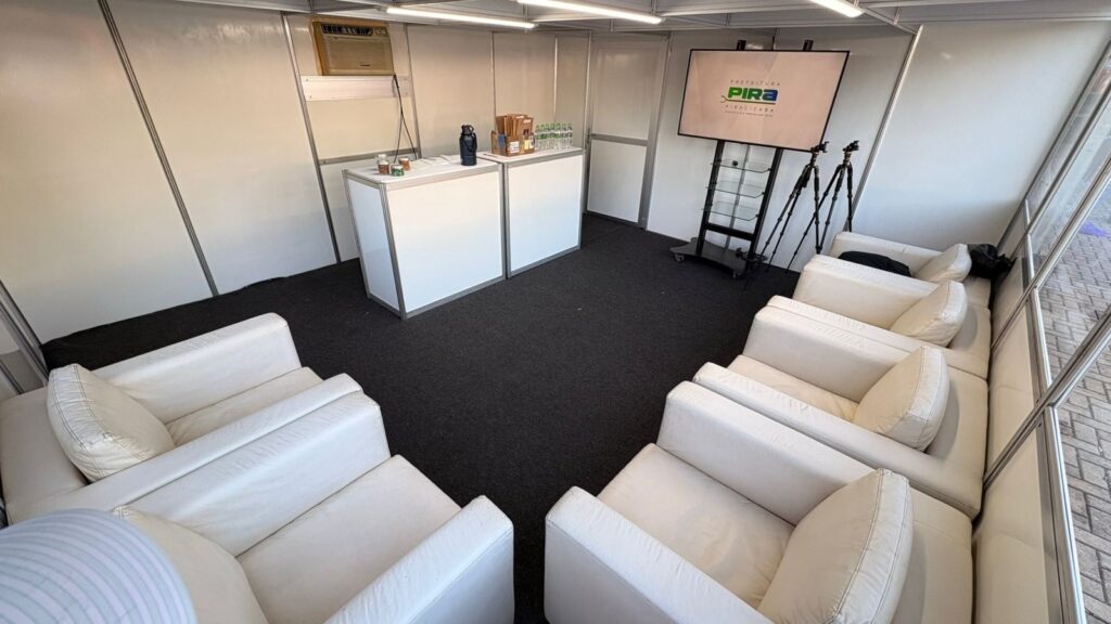 A imagem mostra o interior de uma estrutura temporária, possivelmente um estande de evento, escritório móvel ou espaço de atendimento. As paredes são modulares e brancas, criando um ambiente clean e organizado. O piso é coberto por um carpete escuro, que contrasta com os móveis claros. No centro do espaço, há um conjunto de poltronas brancas dispostas em formato de “U”, sugerindo um ambiente para reunião, conversa ou espera. Na frente dessas poltronas, há duas bancadas brancas alinhadas lado a lado, sobre as quais estão alguns objetos, como garrafas de água, sacolas e outros itens pequenos. À direita da imagem, há uma televisão de tela plana montada sobre um suporte com rodinhas, exibindo o logotipo da “PREFEITURA DE PIRACICABA”. Próximo à TV, há dois tripés, provavelmente para câmeras ou equipamentos de iluminação. A iluminação é feita por lâmpadas lineares no teto, proporcionando um ambiente bem iluminado e funcional. A disposição dos móveis e equipamentos sugere um espaço preparado para recepção, atendimento ao público ou apresentação de informações relacionadas à Prefeitura de Piracicaba.