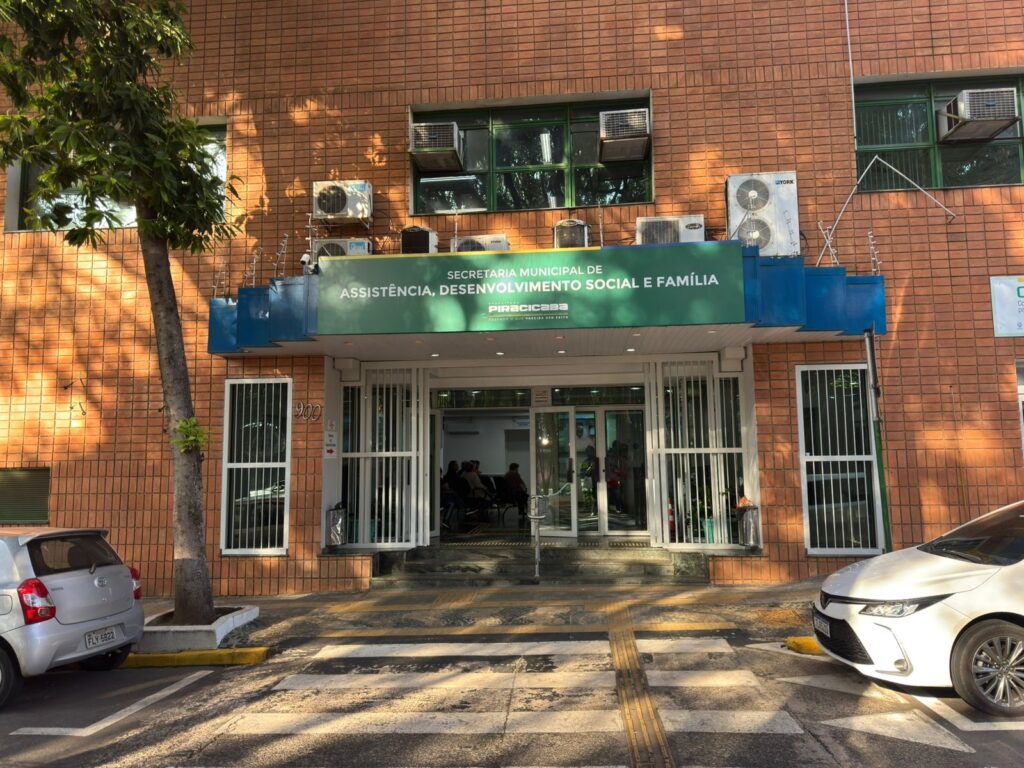 A imagem mostra a fachada de um prédio público, especificamente a Secretaria Municipal de Assistência, Desenvolvimento Social e Família da Prefeitura de Piracicaba. A construção é feita com tijolos aparentes e possui várias unidades de ar-condicionado instaladas acima da entrada principal. A entrada é composta por portas de vidro com grades metálicas, e há uma placa verde com letras brancas indicando o nome da secretaria e da prefeitura, além do slogan "Fazendo o que precisa ser feito". Na calçada em frente ao prédio, há um passeio com piso tátil para acessibilidade, e algumas pessoas estão sentadas no interior da secretaria, visíveis através das portas de vidro. À esquerda da imagem, há uma árvore que fornece sombra, e à direita, um carro branco está estacionado. Também é possível ver parcialmente um carro prata à esquerda da imagem. A cena parece ser durante o dia, com luz solar iluminando a fachada e criando sombras. A imagem transmite a ideia de um serviço público voltado para assistência social, com estrutura adequada para atendimento à população.