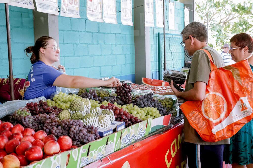 A imagem mostra uma cena cotidiana em uma feira ou mercado de frutas. * À esquerda, uma atendente jovem, usando óculos e uma camiseta azul, está servindo um cliente. Ela estende a mão para entregar um cacho de uvas escuras para um homem que está à sua frente. * O homem, de meia-idade, usa óculos e uma camiseta verde-oliva. Ele segura uma embalagem plástica com uvas e carrega uma sacola de compras laranja grande, da rede Atacadão, sobre o ombro. * Ao lado do homem, uma mulher de cabelos curtos avermelhados observa a transação. * A banca está repleta de frutas frescas, incluindo diversas variedades de uvas (verdes e roxas), maçãs vermelhas em primeiro plano, frutas-do-conde (ou cherimoias) em embalagens de proteção e pedaços de melancia ao fundo. * O ambiente possui paredes de blocos de concreto pintadas de azul claro, e há vários papéis brancos pendurados em um varal acima da banca, possivelmente cartazes ou avisos informativos.