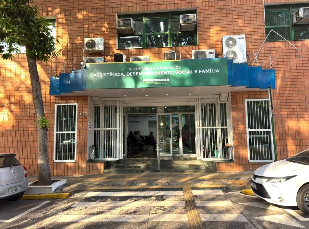 A imagem mostra a fachada de um prédio público, identificado como a "Secretaria Municipal de Assistência, Desenvolvimento Social e Família" da Prefeitura de Piracicaba. O edifício é revestido com azulejos de cor marrom avermelhada. Acima da entrada principal há uma grande placa verde com o nome da secretaria e o logotipo da prefeitura, além do slogan "Fazendo o que precisa ser feito". A entrada é composta por portas de vidro com grades de proteção branca. No interior, é possível ver algumas pessoas sentadas, provavelmente em uma área de espera. Na parte superior da fachada, há várias unidades de ar-condicionado instaladas. Do lado esquerdo da imagem, há uma árvore próxima à calçada, e na rua estão estacionados dois carros, um prata e outro branco. O número "900" está visível na parede ao lado da porta de entrada. No chão, há uma faixa de pedestres com piso tátil para acessibilidade. A iluminação indica que a foto foi tirada durante o dia.