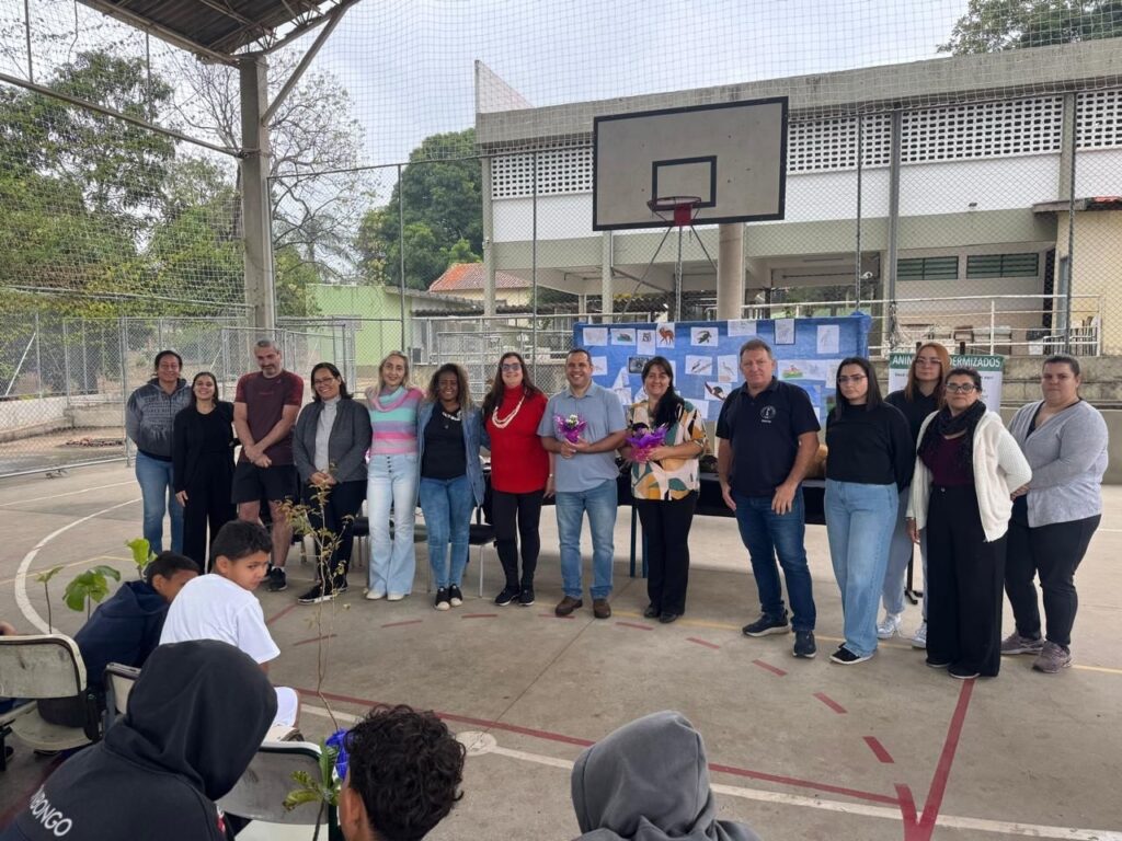 A imagem mostra um grupo de adultos alinhados em um espaço coberto que parece ser uma quadra poliesportiva, provavelmente em uma escola ou centro comunitário. Eles estão de pé, voltados para um grupo de crianças sentadas no chão, que parecem estar assistindo a uma apresentação ou evento. Dois adultos no centro seguram pequenos buquês de flores, sugerindo que podem ter sido homenageados ou que estão celebrando algo. Atrás do grupo há um painel azul com desenhos e fotografias de animais, e uma faixa com o texto "ANIMAIS TAXIDERMIZADOS", indicando que o tema do evento pode estar relacionado à conservação, educação ambiental ou exposição sobre animais empalhados. O ambiente é ao ar livre, mas coberto, com uma cesta de basquete ao fundo e uma cerca de metal, o que reforça a ideia de ser uma quadra esportiva. O público infantil está atento, e a atmosfera parece ser de aprendizado ou celebração escolar.