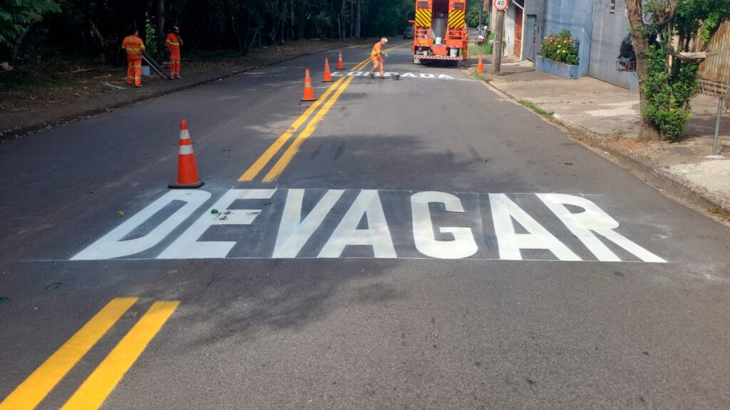 A imagem mostra uma cena de manutenção viária em uma rua asfaltada, provavelmente em uma área urbana ou suburbana. No centro da imagem, está pintada no chão a palavra "DEVAGAR" em letras grandes e brancas, indicando para os motoristas reduzirem a velocidade. Mais adiante, é possível ver outra pintura no asfalto com a palavra "LOMADA", que alerta sobre uma lombada na via. Também há uma placa de limite de velocidade indicando 40 km/h. Na rua, há cones laranjas posicionados ao longo da linha amarela dupla que divide as faixas, sinalizando uma área de trabalho. Três trabalhadores vestidos com uniformes laranjas de alta visibilidade estão presentes, dois próximos à calçada e um perto de um caminhão utilitário laranja estacionado na via, que parece estar envolvido na pintura ou manutenção da estrada. Ao redor, há árvores e vegetação de um lado da rua, e do outro, uma calçada com um muro azul. O dia está claro e ensolarado, com boa iluminação natural. A cena transmite a ideia de um trabalho de pintura e sinalização viária em andamento, com medidas para garantir a segurança dos trabalhadores e dos motoristas.