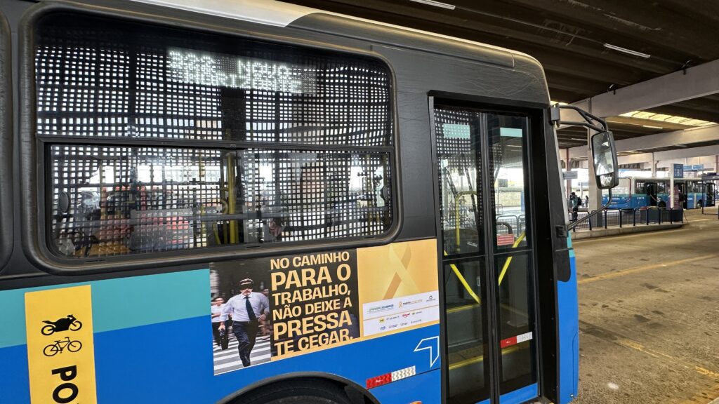 A imagem mostra um ônibus urbano estacionado em um terminal coberto. O ônibus é azul e preto, com um grande adesivo publicitário na lateral. O adesivo exibe a imagem de um homem vendado atravessando uma faixa de pedestres, transmitindo uma mensagem de conscientização sobre segurança no trânsito. O texto do adesivo diz: "NO CAMINHO PARA O TRABALHO, NÃO DEIXE A PRESSA TE CEGAR", sugerindo que as pessoas devem evitar a pressa para prevenir acidentes. Na lateral do ônibus, há também um adesivo amarelo com símbolos de motocicleta e bicicleta, indicando que o veículo pode ter algum tipo de relação ou espaço para esses meios de transporte, ou que a campanha abrange motoristas e ciclistas. O terminal onde o ônibus está estacionado possui estrutura de concreto com iluminação artificial e vigas no teto. Ao fundo, é possível ver outro ônibus azul estacionado. A cena parece ser durante o dia, mas em ambiente coberto, com luz difusa. A imagem transmite uma mensagem de segurança no trânsito e prevenção de acidentes, focando na importância de não se apressar no caminho para o trabalho.