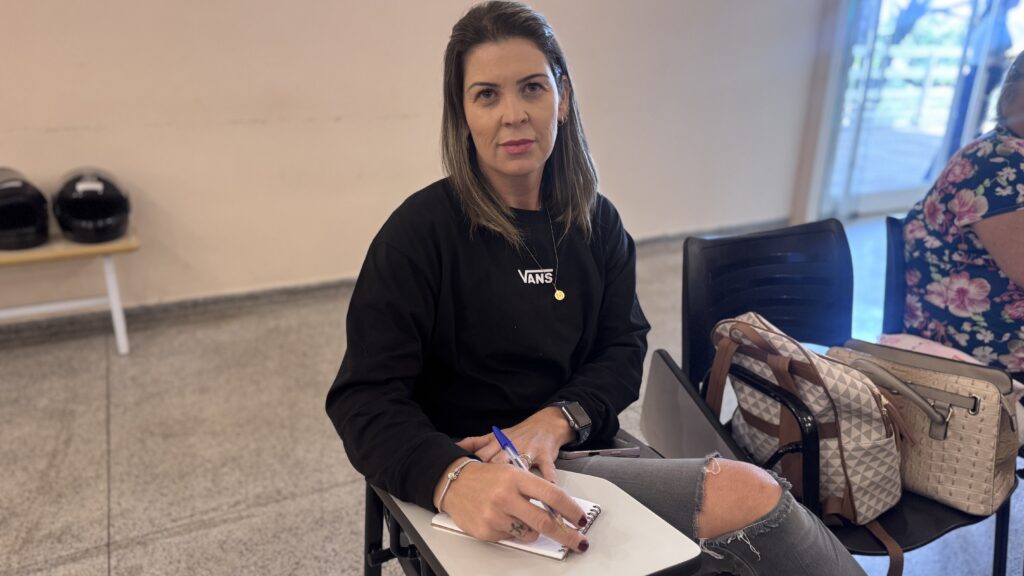 A imagem mostra uma mulher sentada em uma sala de aula ou auditório, em uma cadeira com uma pequena mesa acoplada onde ela está escrevendo em um caderno com uma caneta azul. Ela tem cabelo castanho na altura dos ombros, com mechas mais claras, e usa um moletom preto com o logo "VANS" na frente. Está vestindo também uma calça jeans cinza com rasgo no joelho. Ela usa um relógio no pulso esquerdo e um colar delicado. Ao lado dela, sobre um banco branco, estão dois capacetes pretos, provavelmente de motocicleta. Ao fundo, há outra pessoa parcialmente visível, vestindo uma blusa com estampa floral e com uma bolsa estampada pendurada na cadeira. A iluminação é suave e natural, e o ambiente parece ser uma sala de aula moderna e simples, com paredes claras e uma porta visível ao fundo. A mulher está olhando diretamente para a câmera, com uma expressão neutra.