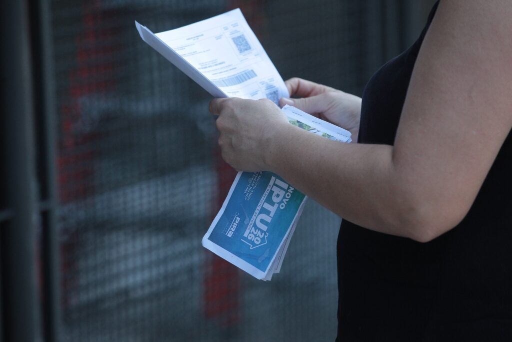 A imagem mostra uma pessoa vista de perfil, vestindo uma blusa preta, segurando um conjunto de documentos em suas mãos. * O destaque principal é um folheto azul com o texto "NOVO IPTU 26", que inclui o logotipo da prefeitura de Piracicaba e a frase "O BENEFÍCIO É DE TODOS". * Acima desse folheto, a pessoa segura uma guia de pagamento oficial, onde é possível notar um código de barras e um código QR, elementos típicos de boletos bancários brasileiros. * O fundo da imagem é composto por uma grade metálica cinza, mantendo o foco total na pessoa e nos documentos que ela está manuseando.