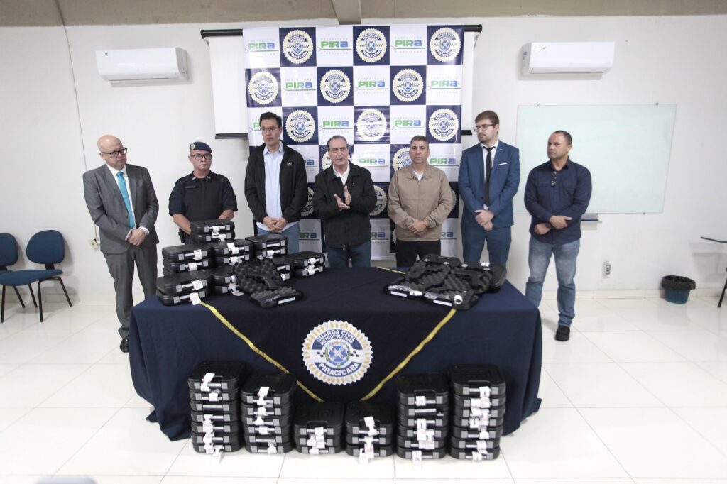 A imagem mostra um evento oficial, possivelmente uma cerimônia de entrega ou apresentação de equipamentos para a Guarda Civil Metropolitana de Piracicaba. Sete homens estão alinhados atrás de uma mesa coberta por um pano azul escuro, com o logotipo da Guarda Civil Metropolitana de Piracicaba estampado na frente. Sobre a mesa e no chão à frente dela, há várias caixas rígidas pretas, algumas abertas, exibindo armas de fogo. Os homens estão vestidos de forma variada: alguns usam ternos sociais, outros roupas mais casuais, e um deles está com uniforme policial completo, incluindo um boina preta. O fundo da sala é uma parede branca com um painel repetitivo contendo os logotipos da Prefeitura de Piracicaba e da Guarda Civil. A sala é bem iluminada, com um piso claro e duas unidades de ar-condicionado instaladas na parede. A disposição dos participantes e dos objetos transmite um clima formal e organizado.