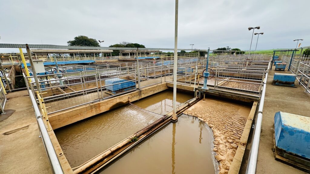 A imagem mostra uma estação de tratamento de águas residuais industriais, com vários tanques retangulares de decantação. A água nos tanques está turva e de cor marrom, indicando a presença de resíduos e sedimentos. Em um dos tanques, é possível ver uma camada espessa de lodo acumulado na superfície. Estruturas metálicas como corrimãos e passarelas conectam os tanques, facilitando o acesso e a manutenção do sistema. Há tubulações visíveis, incluindo uma que despeja água em um dos tanques, criando ondulações na superfície. O ambiente é industrial, com iluminação natural em um dia nublado, e ao fundo aparecem árvores e áreas abertas, sugerindo que a estação está localizada em uma região rural ou semi-rural. A imagem transmite uma sensação funcional e utilitária, típica de instalações de tratamento de água.