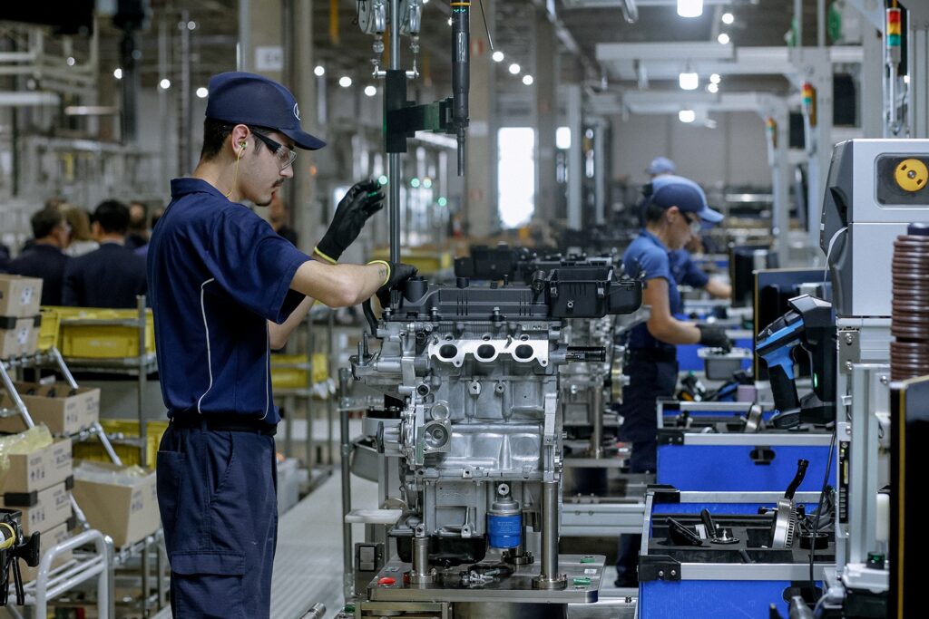 A imagem mostra uma linha de montagem automotiva, com foco em um trabalhador que está montando um motor. O motor é o elemento principal da foto, visível com várias peças metálicas, como a cabeça do cilindro, filtro de óleo e diversos tubos e fios. O trabalhador, à esquerda, veste um uniforme azul escuro, boné com um logo discreto, óculos de proteção e luvas pretas, concentrado na montagem do motor. A iluminação é forte e típica de ambiente industrial, destacando os detalhes metálicos do motor e das máquinas ao redor. Ao fundo, outros trabalhadores, com uniformes semelhantes, também estão envolvidos no processo de montagem, indicando uma linha de produção movimentada. A foto tem um foco nítido no motor e no trabalhador, enquanto o fundo está um pouco desfocado, o que ajuda a destacar a complexidade do trabalho. As cores predominantes são tons frios dos metais e uniformes, com pequenos detalhes coloridos em luzes de advertência e etiquetas. A perspectiva é levemente inclinada, mostrando bem as peças do motor e as ações do trabalhador. A imagem transmite uma sensação de precisão, esforço e produção industrial.