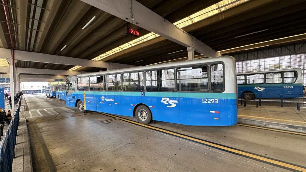 A imagem mostra um terminal de ônibus urbano, provavelmente na cidade de Piracicaba, Brasil. No primeiro plano, há um ônibus azul e prata com o número de frota 12293 e o brasão da cidade de Piracicaba na lateral, indicando que ele pertence à empresa de transporte público local. Outros ônibus podem ser vistos estacionados ao fundo, incluindo um com o número 32813. O terminal possui um teto de concreto com iluminação embutida e um relógio digital que marca 12:06. O piso é de concreto e há faixas amarelas que delimitam as áreas de embarque e desembarque dos passageiros. A cena é iluminada pela luz natural do dia, transmitindo uma atmosfera funcional e típica de um ponto de transporte público em funcionamento.