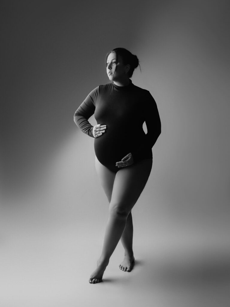 A imagem é uma fotografia em preto e branco de uma mulher grávida, capturada em estúdio. Ela está em pé, com as pernas cruzadas, segurando suavemente sua barriga de gestante com as duas mãos. Seu olhar está direcionado para o lado esquerdo, transmitindo uma expressão contemplativa. A mulher veste um body justo de manga longa e gola alta, que destaca a forma de seu corpo e a barriga da gravidez. Seu cabelo está preso em um coque, e ela está descalça. A iluminação é suave e dramática, vindo do lado esquerdo, criando um efeito de luz e sombra que realça suas formas e dá profundidade à imagem. O fundo é simples, em tons de cinza, sem elementos que distraiam, mantendo o foco totalmente na mulher. A fotografia tem uma textura granulada que contribui para um aspecto artístico e elegante. O clima geral da imagem é sereno e sofisticado.