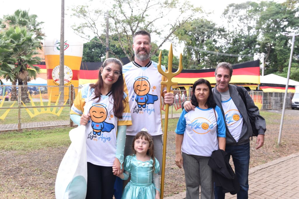 A imagem mostra um grupo de cinco pessoas posando para uma foto ao ar livre, provavelmente durante um evento cultural ou comunitário. No centro, há um homem alto com barba, vestido com uma camiseta branca e amarela da "Liga dos Anjos", segurando um tridente dourado. Ao lado dele, à esquerda, está uma mulher com cabelos longos e escuros, usando óculos e uma camiseta da mesma liga. Na frente deles, uma menina pequena usa um vestido azul claro de princesa e uma tiara na cabeça. À direita do homem de camiseta amarela, há uma mulher mais velha vestindo uma camiseta branca com a palavra "Anjos" e calças cinza estampadas, e ao lado dela, um homem com camiseta branca, jeans escuros e jaqueta cinza. Ao fundo, há uma grande estrutura inflável em forma de copo de cerveja, árvores e faixas com as cores da bandeira alemã (preto, vermelho e amarelo), sugerindo que o evento pode estar relacionado a uma celebração típica alemã, como a Oktoberfest. A iluminação é natural, indicando que a foto foi tirada durante o dia, e o grupo está posicionado em um caminho pavimentado em frente a uma cerca de arame.