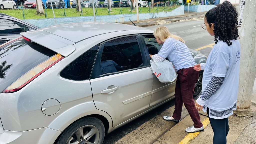A imagem mostra uma cena ao ar livre, em um dia nublado, onde duas mulheres estão interagindo com o motorista de um carro prata parado na beira da estrada. * A mulher à esquerda, de cabelos loiros, está inclinada para dentro da janela do passageiro do veículo, entregando uma sacola plástica branca. Ela veste uma blusa branca de manga comprida e calças vinho. * Ao lado dela, à direita, uma mulher de cabelos cacheados escuros observa a interação. Ela usa uma camiseta branca com estampas nas costas, mangas que parecem ter um padrão xadrez por baixo e calças pretas. * O carro prata é um modelo Ford Focus hatch. * Ao fundo, vê-se uma área gramada, uma cerca de proteção, um poste de concreto e outros veículos estacionados ou circulando ao longe, sugerindo um ambiente urbano ou uma via de acesso. O chão parece estar úmido, indicando que pode ter chovido recentemente.
