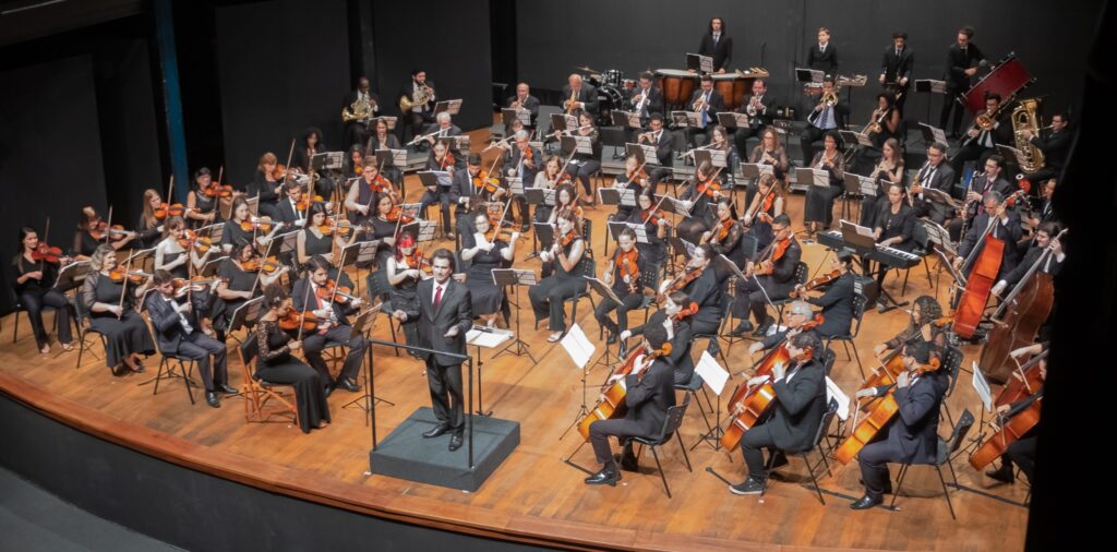 A imagem mostra uma orquestra sinfônica completa em apresentação, posicionada em um palco de madeira. O maestro está no centro, de costas para o público, conduzindo os músicos com os braços levemente elevados. Os integrantes da orquestra, vestidos majoritariamente com roupas formais pretas, estão organizados em fileiras curvas ao redor do maestro. Vários instrumentos são visíveis, incluindo cordas (violinos, violas, violoncelos e contrabaixos), metais (trompas, trompetes, trombones, tubas), madeiras e percussão. A iluminação está focada nos músicos, destacando o brilho dos instrumentos e o piso de madeira, enquanto o fundo permanece escuro, criando um ambiente intimista e concentrado na performance artística. A foto foi tirada de um ângulo elevado, proporcionando uma visão ampla e detalhada da orquestra em ação.