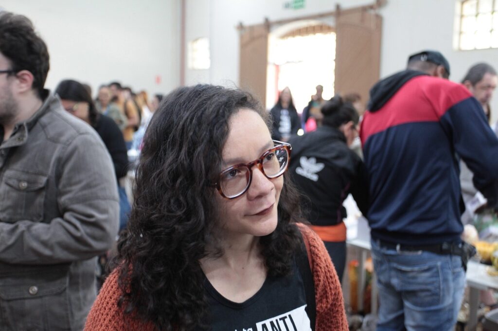 Esta imagem captura uma cena em um ambiente interno, possivelmente um evento ou feira, com diversas pessoas ao fundo. * O foco principal é uma mulher em primeiro plano, posicionada ligeiramente à direita do centro. Ela tem cabelos longos, escuros e cacheados, usa óculos de armação marrom e veste uma camiseta preta com a inscrição "ANTI" parcialmente visível, coberta por um cardigã ou suéter de cor ferrugem ou laranja queimado. Ela olha para a direita com uma expressão serena. * À esquerda, vemos o perfil de um homem usando uma jaqueta cinza texturizada. * Ao fundo, há várias outras pessoas em diferentes planos, criando uma sensação de um local movimentado. Vê-se uma pessoa vestindo uma jaqueta preta com o logotipo da Adidas, e outra pessoa à direita com uma jaqueta de cores azul e vermelha. O ambiente possui iluminação natural vinda de grandes aberturas ou janelas ao fundo, que parecem ser portas de madeira.