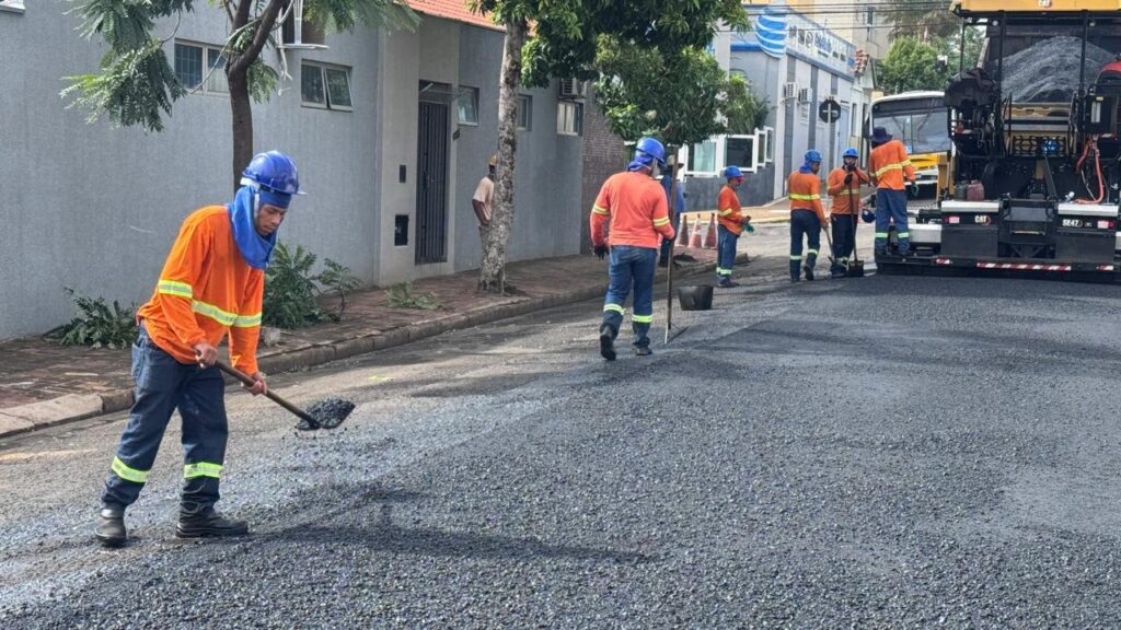A imagem mostra uma equipe de trabalhadores realizando obras em uma rua durante o dia. No centro da imagem, um trabalhador está espalhando asfalto com uma pá, enquanto outros, todos vestidos com camisas laranja de manga longa, calças jeans e capacetes azuis, trabalham ao redor. À direita, há uma máquina grande e amarela da marca CAT, usada para pavimentação. A rua está parcialmente bloqueada para o serviço, e ao fundo é possível ver prédios cinzas e algumas árvores. O ambiente está iluminado pela luz natural do sol, indicando que o trabalho está acontecendo em um dia claro. A cena transmite a ideia de esforço e trabalho em equipe na manutenção ou construção da via.