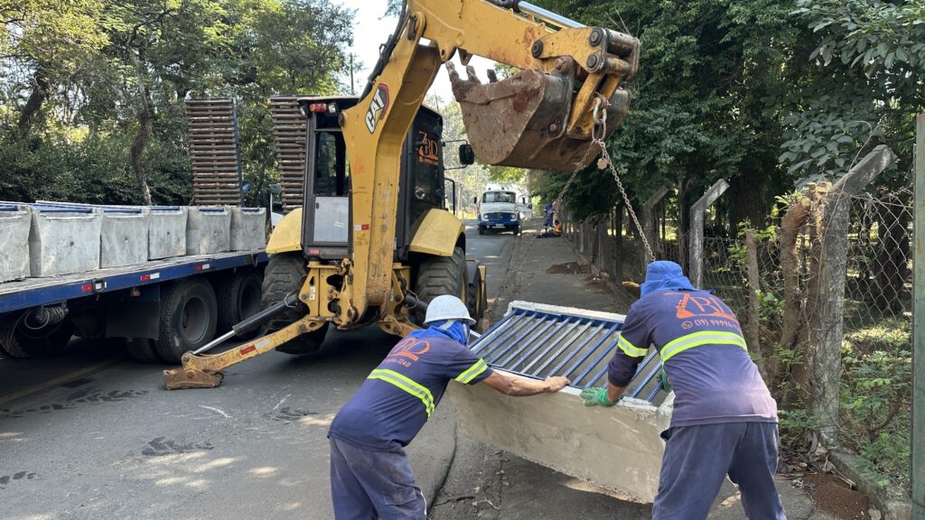 Maquinário: retroescavadeira amarela (Caterpillar) levantando uma peça pré-moldada de concreto com corrente. Ação: dois operários da RD Construções (uniformes azul‑marinho com faixas refletivas) guiam e posicionam a peça — parece instalação de bueiro/sistema de drenagem com grade metálica azul no topo. Transporte e apoio: caminhão à esquerda carregado com peças similares; outros funcionários ao fundo auxiliando ou observando. Ambiente: via pública durante o dia, com vegetação ao fundo e área de trabalho delimitada por cerca de arame.