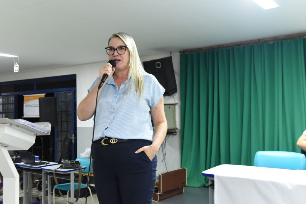 A imagem mostra uma mulher loira falando ao microfone em um ambiente interno que parece ser um auditório ou sala de eventos, possivelmente em uma escola ou centro comunitário. Ela usa uma camisa azul clara de manga curta, calças escuras e óculos de grau com armação preta. A mulher está em pé, em frente a um púlpito de madeira, segurando o microfone com uma mão, enquanto a outra está no bolso da calça. Atrás dela, há uma cortina verde e uma mesa com um notebook e um cartaz pequeno, que tem o texto "vagas em aberto". A iluminação é clara, e o ambiente parece preparado para uma apresentação ou palestra. A expressão da mulher é séria e concentrada, indicando que ela está falando sobre um assunto importante.