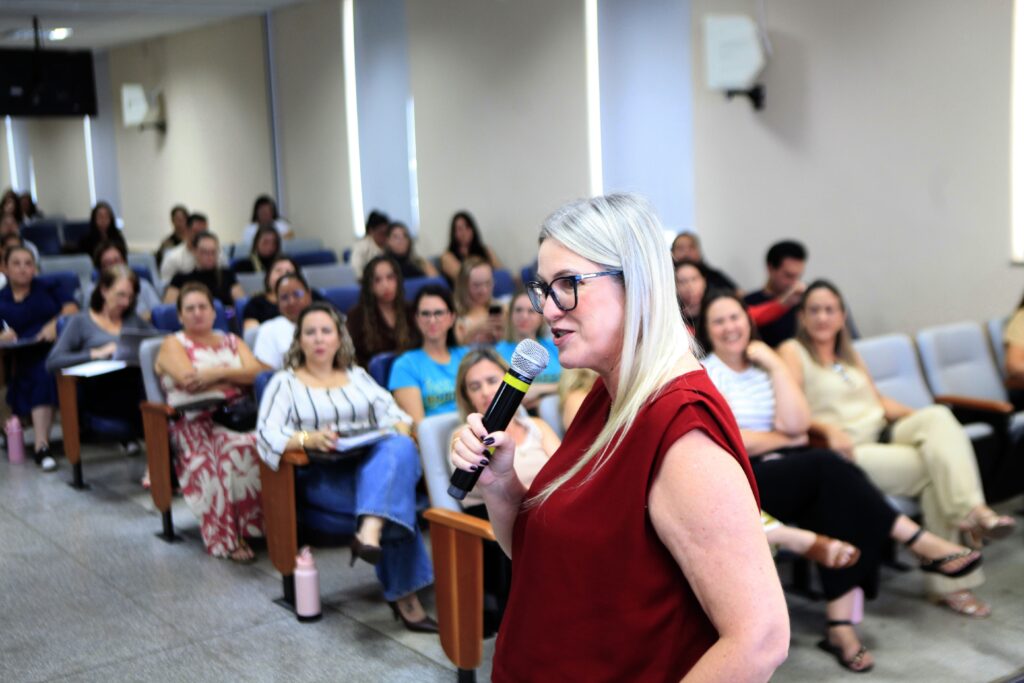 A imagem mostra uma mulher falando ao microfone em um ambiente que parece ser uma sala de palestras ou conferências. Ela está em primeiro plano, um pouco à direita. A mulher tem cabelo loiro, usa óculos e veste uma blusa sem mangas vermelha escura. Ela parece estar falando com entusiasmo ou explicando algo. Ao fundo, há várias pessoas sentadas em cadeiras azuis, formando uma audiência. A maioria das pessoas na audiência são mulheres, mas seus rostos estão desfocados, o que dá destaque à mulher que está falando. O ambiente parece profissional, com paredes claras e iluminação artificial uniforme. É um momento típico de apresentação ou palestra, com a oradora dirigindo-se ao público.