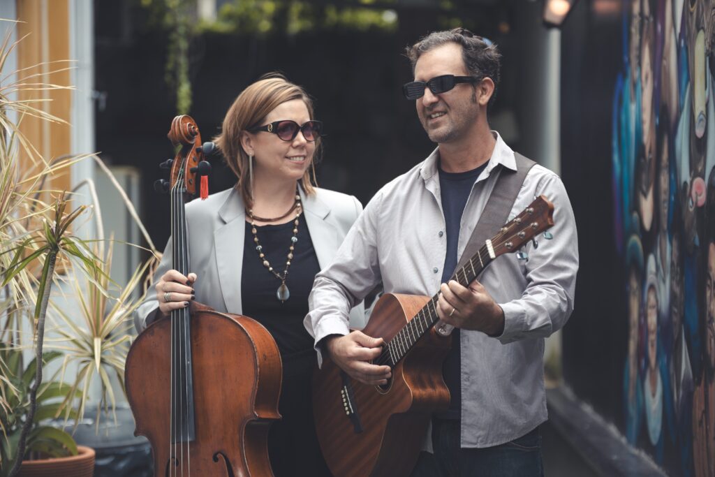 A imagem mostra um dueto musical posicionado ao ar livre em um ambiente urbano. * À esquerda, uma mulher de cabelos curtos, vestindo um blazer cinza sobre uma blusa preta, segura um violoncelo de madeira envelhecida. Ela usa óculos escuros e um colar longo com um pingente. * À direita, um homem também de óculos escuros, vestindo uma camisa social de botões listrada sobre uma camiseta escura, toca um violão acústico. Ele olha para a mulher enquanto ela olha para ele, sugerindo uma interação musical. * O fundo apresenta uma parede com um mural artístico colorido e plantas em um vaso à esquerda, criando um contraste interessante entre a sofisticação dos músicos e o cenário urbano e criativo. A iluminação é suave e natural.