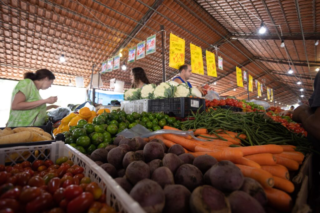 A imagem retrata uma cena movimentada em uma feira livre coberta, com uma grande variedade de produtos frescos em destaque. * Em primeiro plano, há caixas plásticas organizadas contendo diversos vegetais, como beterrabas, cenouras, tomates, pimentões amarelos, vagens e pequenos frutos verdes, provavelmente jilós ou berinjelas. * Ao fundo, é possível ver algumas pessoas, incluindo uma mulher de blusa verde à esquerda e um homem ao centro, que parece estar atendendo ou organizando a mercadoria perto de caixas de couve-flor. * O ambiente possui um teto de telhas de barro, e há várias placas amarelas penduradas com preços escritos à mão, indicando valores para itens como tomate e limão, o que confere um aspecto típico de mercado local. * A iluminação é composta por lâmpadas pendentes que iluminam as bancadas, criando um contraste entre as cores vibrantes dos vegetais e a estrutura rústica do local.