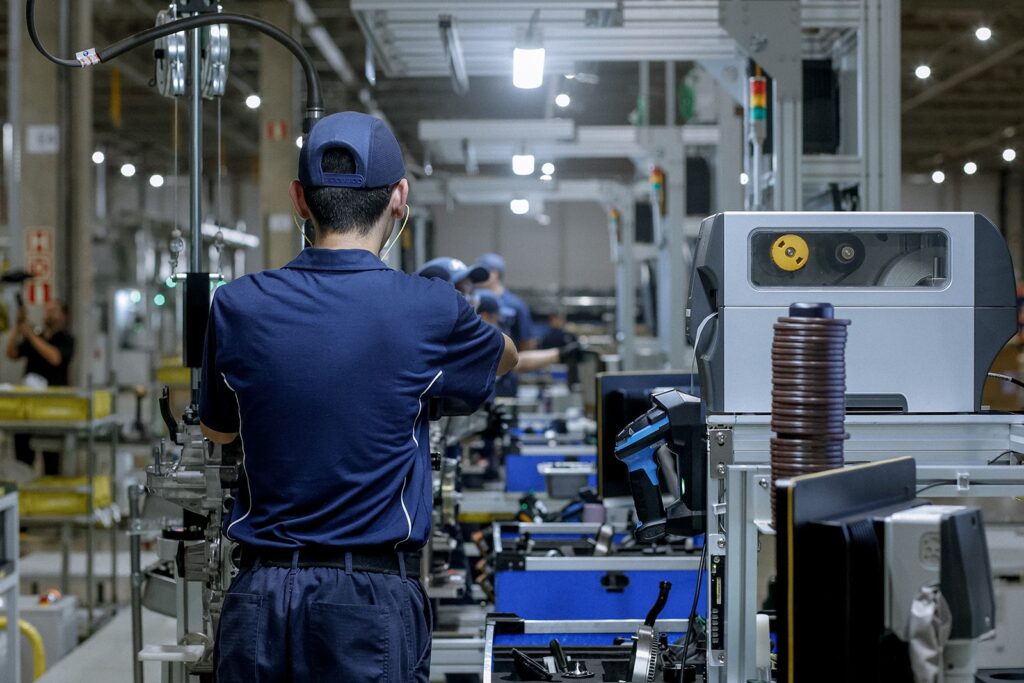 A imagem retrata um ambiente de linha de montagem industrial. Em primeiro plano, vemos um funcionário de costas, vestindo uma camisa polo azul e boné, operando um equipamento em uma estação de trabalho. * O trabalhador está focado em sua tarefa, cercado por componentes mecânicos e ferramentas. * À direita, há uma máquina de etiquetagem ou marcação, com uma pilha de componentes circulares empilhados ao lado. * Ao fundo, a perspectiva da fábrica revela outros trabalhadores em suas respectivas estações, indicando um processo produtivo em larga escala. * O ambiente é iluminado por luzes artificiais frias, típicas de instalações industriais, e apresenta uma organização estruturada de equipamentos, caixas azuis e componentes metálicos.