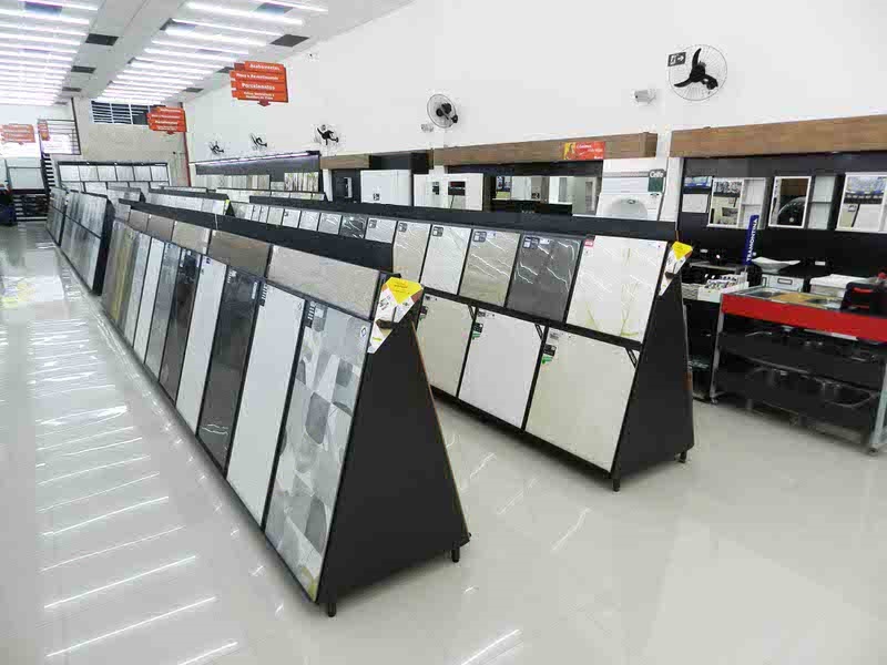 A imagem mostra o interior de uma loja de materiais para construção ou decoração, especializada em revestimentos, como pisos e azulejos. No centro da imagem, há duas estruturas em formato de cavalete, dispostas em fileiras paralelas, que exibem diversos modelos de pisos cerâmicos ou porcelanatos. Esses revestimentos apresentam diferentes padrões, cores e texturas, incluindo opções lisas em tons de branco e cinza, além de alguns com desenhos decorativos. O piso da loja é feito de azulejos claros e brilhantes, que refletem a iluminação forte e uniforme vinda das lâmpadas fluorescentes no teto. Ao fundo, é possível ver outras amostras e displays montados nas paredes, além de uma bancada com pia, que tem um adesivo da marca "Tramontina". No teto, há placas laranjas que provavelmente indicam setores ou categorias de produtos. O ambiente está limpo, organizado e bem iluminado, típico de uma área de exposição para clientes escolherem os revestimentos adequados para suas reformas ou construções. Não há pessoas na imagem, apenas os produtos em exposição.