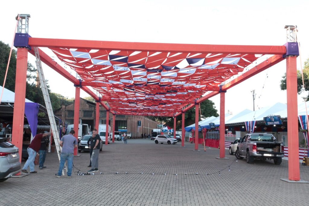 A imagem mostra uma área externa pavimentada, provavelmente um pátio ou estacionamento, onde está montada uma estrutura grande e retangular com uma moldura vermelha sustentada por quatro pilares verticais. O teto dessa estrutura é coberto por uma lona decorativa em tons de vermelho, branco e azul escuro, formando um padrão geométrico que lembra uma rede. Pendentes sob o teto, há fileiras de pequenas lâmpadas redondas, indicando que o local pode ser iluminado à noite. Na parte frontal da imagem, três homens estão trabalhando: dois deles manipulam um escada metálica alta e fios elétricos, enquanto o terceiro observa. Ao redor da estrutura, há vários veículos estacionados, incluindo carros e vans. À direita, há tendas brancas temporárias, uma das quais exibe a inscrição "ESTADOS UNIDOS", sugerindo que a área pode ser destinada a algum tipo de evento cultural, feira ou exposição relacionada aos Estados Unidos. Ao fundo, vê-se um conjunto de edifícios de tijolos, possivelmente antigas construções industriais, que dão um ar urbano e histórico ao local. A iluminação natural indica que a foto foi tirada durante o dia. A cena transmite a ideia de preparação para um evento ao ar livre, com atenção aos detalhes da decoração e infraestrutura.