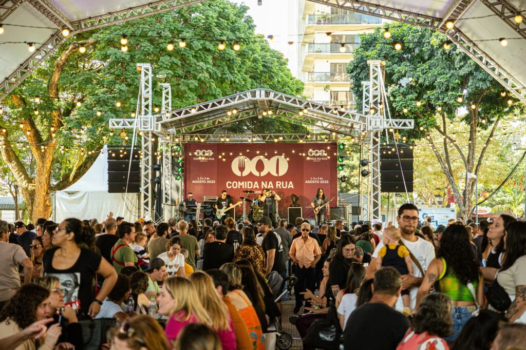A imagem mostra um evento ao ar livre, provavelmente um festival de comida e música, realizado sob uma grande tenda decorada com luzes de cordão. No centro da cena, há um palco onde uma banda está se apresentando, com instrumentos e equipamentos de som visíveis. Atrás do palco, há uma grande faixa vermelha com o texto "COMIDA NA RUA" e as datas "1, 2 e 3 de AGOSTO 2025", indicando o nome e a data do evento. Na frente do palco, uma multidão diversificada de pessoas está reunida, algumas em pé e outras sentadas em mesas, apreciando a música e o ambiente. A iluminação é uma combinação de luz natural do dia e luzes artificiais, criando uma atmosfera acolhedora e animada. No fundo, é possível ver árvores e um edifício de vários andares, sugerindo que o evento acontece em um parque ou praça urbana. A imagem transmite uma sensação de comunidade, cultura e celebração em um cenário vibrante e festivo.