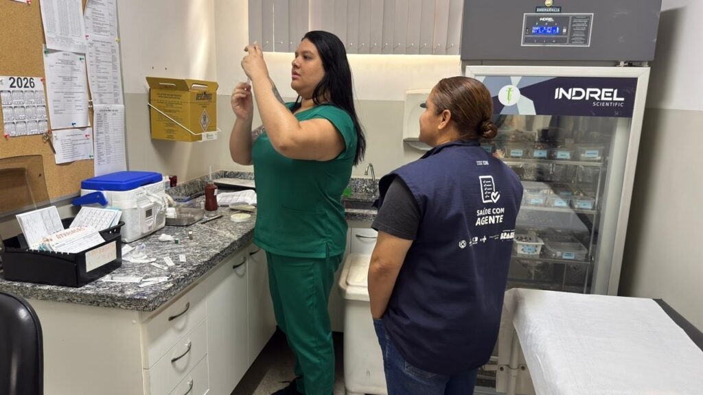 A imagem mostra um ambiente clínico ou laboratório médico, onde duas pessoas estão envolvidas em um procedimento relacionado à saúde. Uma delas, vestindo uniforme verde, está preparando uma seringa, possivelmente para uma vacinação ou coleta de amostra. A outra pessoa, usando colete escuro com inscrições que parecem indicar que é um agente de saúde, observa atentamente. No fundo, há um refrigerador científico de grande porte, provavelmente utilizado para armazenar vacinas ou amostras médicas, com várias gavetas transparentes contendo materiais. À esquerda, um quadro de avisos exibe um calendário do ano de 2026 e alguns documentos. Em cima de uma bancada de granito, estão dispostos vários materiais médicos, como seringas, frascos e bandejas metálicas. O ambiente é bem iluminado, com paredes azuis e brancas e armários, sugerindo um local organizado e preparado para procedimentos médicos. Ao centro, a interação entre os profissionais evidencia um momento de cuidado e atenção na área da saúde.