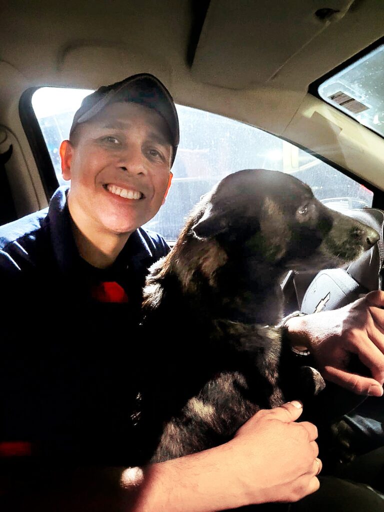 A imagem mostra um homem e seu cachorro dentro de um carro. O homem está sorrindo para a câmera, usando uma camisa polo azul escura e um boné. Ele parece estar feliz e relaxado. O cachorro, que tem pelagem escura, está ao lado dele, olhando para fora da janela do carro. O volante do carro, com o logo da Chevrolet, também é visível. A luz do sol entra pela janela, iluminando o rosto do homem e parte do cachorro, criando um ambiente acolhedor e alegre. A cena transmite uma sensação de companheirismo e tranquilidade durante uma viagem ou passeio de carro.