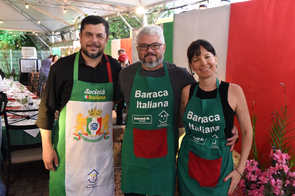 A imagem mostra três pessoas sorridentes, posicionadas lado a lado em um ambiente externo, provavelmente em um evento cultural italiano ou uma barraca de comida típica. À esquerda, há um homem com cabelos escuros e barba, vestindo uma camiseta preta e um avental verde com detalhes em branco e vermelho, onde se lê "Società Italiana". No centro, um homem de cabelos grisalhos e barba, usando óculos, camiseta cinza e avental verde com bolso vermelho, com inscrições "Barraca Italiana", "Unimed Piracicaba" e "Casa do Bom Menino". À direita, uma mulher de cabelos escuros, vestindo regata preta e avental semelhante ao do homem do centro, com as mesmas inscrições. Ao fundo, vê-se uma mesa posta com toalhas brancas e copos, e uma grande bandeira italiana (verde, branco e vermelho) como decoração, além de flores cor-de-rosa artificiais à direita. A iluminação é quente e sugere que o evento ocorre ao entardecer ou à noite. A atmosfera parece alegre e acolhedora, típica de uma festa ou feira temática italiana.