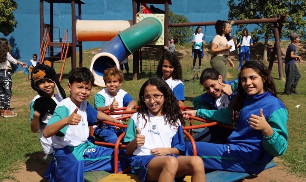 A imagem mostra um grupo de crianças e adolescentes em um parquinho ao ar livre, em um dia ensolarado. Cinco crianças estão sentadas em um brinquedo giratório, todas usando uniformes escolares brancos com detalhes em azul e verde, e parecem felizes, algumas fazendo sinal de positivo com o polegar. Uma das meninas usa óculos, e uma criança à esquerda usa um chapéu de pelúcia que lembra um urso. Ao fundo, há uma estrutura de playground com escorregadores nas cores azul e verde. Outras pessoas, possivelmente adultos ou adolescentes mais velhos, estão em pé ou caminhando no gramado ao redor. O ambiente transmite uma sensação de diversão, amizade e descontração típica de momentos de recreio ou passeio escolar. A palavra "Piracicaba" no uniforme indica possivelmente a cidade ou a instituição escolar.