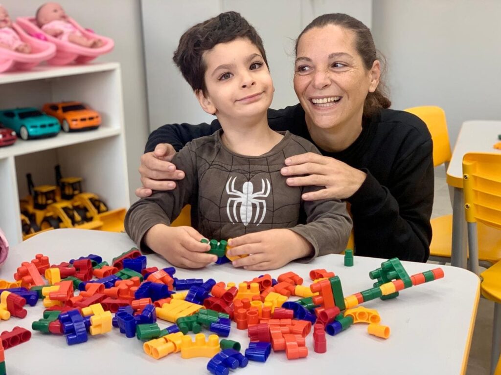 A imagem mostra uma cena afetuosa em um ambiente que parece ser uma sala de terapia infantil ou uma sala de aula. * No centro, uma mulher adulta com um sorriso largo abraça carinhosamente um menino que está sentado à mesa. O menino veste uma camiseta marrom com o desenho do Homem-Aranha e está concentrado, brincando com blocos de montar coloridos. * Sobre a mesa, há uma grande quantidade de peças de montar de plástico nas cores vermelho, azul, amarelo e verde, espalhadas em uma desordem lúdica. * Ao fundo, à esquerda, observa-se uma estante branca com prateleiras organizadas. Na prateleira superior, há dois brinquedos de bebês em seus berços cor-de-rosa; abaixo, encontram-se carrinhos de brinquedo e, na base, veículos de construção amarelos. * O ambiente é iluminado e apresenta mobiliário escolar, como cadeiras amarelas, reforçando a ideia de um espaço dedicado ao desenvolvimento e ao aprendizado infantil.