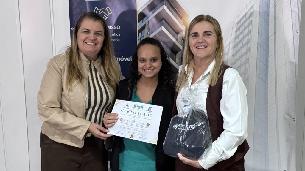 A imagem mostra três mulheres posando para uma foto em um ambiente interno, provavelmente em um evento de entrega de certificados. * A mulher à esquerda veste uma jaqueta bege sobre uma blusa listrada e calça marrom. * No centro, uma jovem com cabelos cacheados escuros veste uma blusa verde e segura um certificado de conclusão do "Curso de Donuts", emitido pela Prefeitura de Piracicaba em parceria com a Ranking Construtora. * À direita, a terceira mulher veste uma camisa branca sob um colete marrom e segura uma sacola de presente com a logomarca da "Ranking Construtora". Ao fundo, há um banner institucional com a imagem de um prédio moderno, reforçando a parceria entre o setor público e a empresa privada na realização do curso profissionalizante. Todas sorriem para a câmera, transmitindo um clima de conquista e celebração.