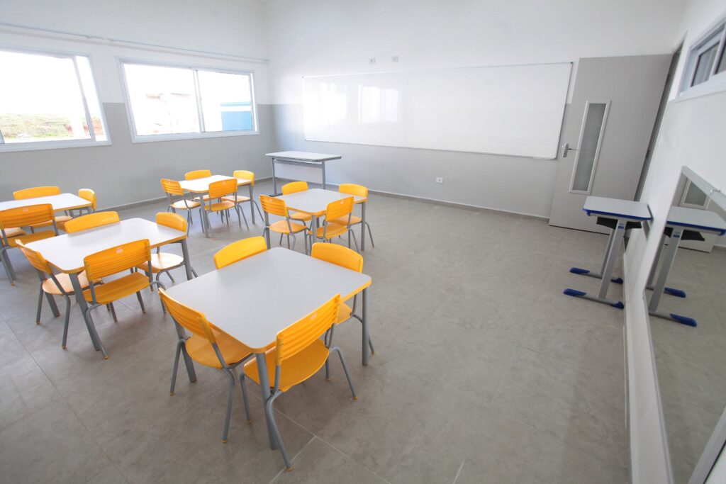 A imagem mostra uma sala de aula moderna e vazia, vista de um ângulo elevado e amplo. A sala é bem iluminada pela luz natural que entra por duas grandes janelas na parede esquerda. As paredes são pintadas de cinza claro, com o teto branco. O piso é composto por grandes azulejos quadrados em tom cinza claro. No ambiente, há várias mesas quadradas cinza, cada uma acompanhada por quatro cadeiras amarelas de plástico, dispostas em um padrão organizado por toda a sala. Na parede da frente, há um grande quadro branco horizontal. À direita, há uma porta cinza com um painel de vidro vertical. Também na parede direita, há uma mesa maior, de cor cinza com detalhes em azul, possivelmente para o professor, e um espelho que reflete parte da mesa. O estilo da sala é minimalista, limpo e funcional, típico de um ambiente educacional moderno. Não há pessoas presentes na imagem.
