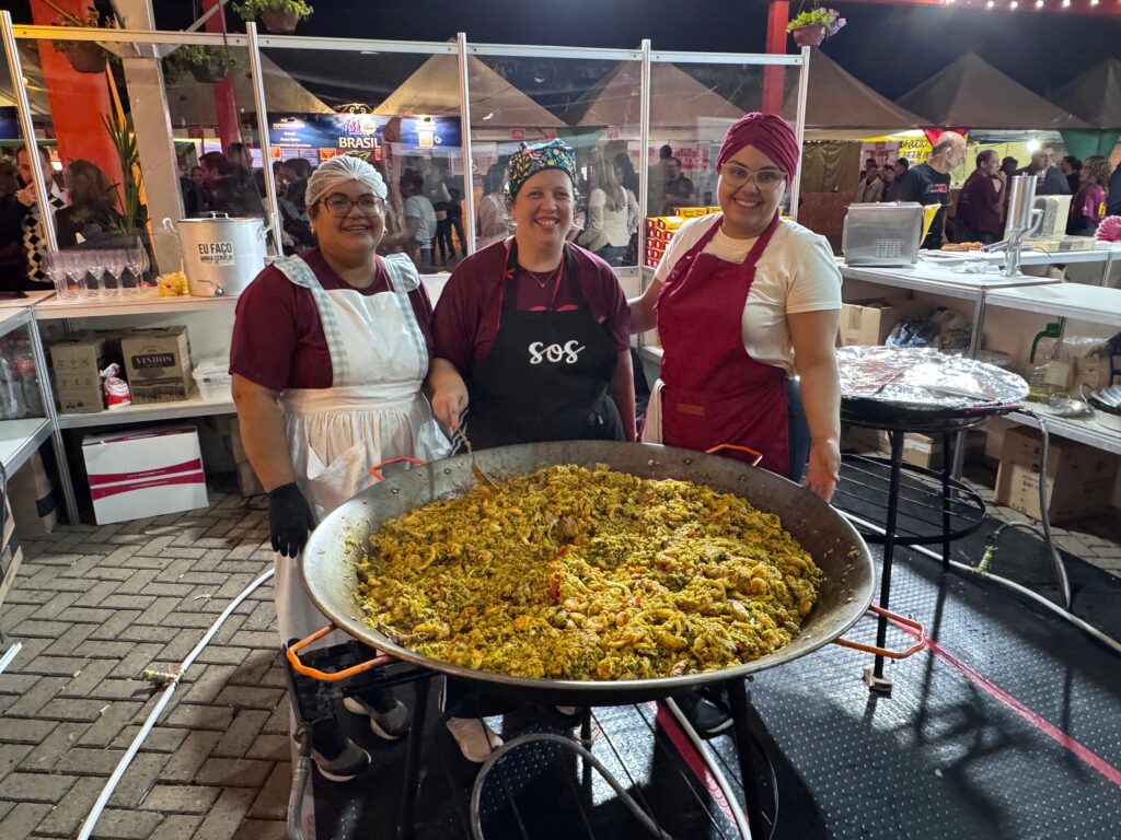 A imagem mostra três mulheres sorridentes em um ambiente ao ar livre, possivelmente em uma feira ou festival noturno de comida. Elas estão posicionadas atrás de uma grande panela de paella cheia de arroz amarelo com frutos do mar. As mulheres vestem uniformes compostos por aventais e lenços ou toucas na cabeça, sugerindo que estão envolvidas na preparação e venda da comida. O cenário é iluminado por luzes artificiais, destacando a barraca onde estão trabalhando, que possui uma bancada de metal e alguns utensílios e caixas ao redor. Ao fundo, é possível ver outras barracas e uma estrutura de lona típica de feiras. No centro da imagem, uma das mulheres usa uma faixa na cabeça com flores, e outra tem um lenço na cabeça, enquanto a terceira usa uma touca branca. Na bancada, há uma grande panela com a inscrição "EU FAÇO MINHA CERVEJA" e caixas com produtos, indicando que também podem estar vendendo bebidas. O ambiente transmite uma atmosfera calorosa e alegre, típica de eventos comunitários ou festivais gastronômicos.