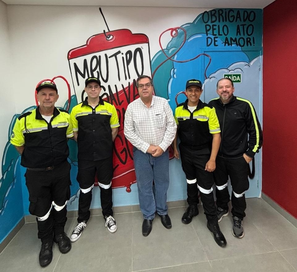 A imagem mostra um grupo de cinco homens posando para uma foto em um ambiente interno, em frente a uma parede decorada com um mural artístico. * O mural, pintado em tons de azul, vermelho e branco, apresenta a frase "MEU TIPO SALVA" dentro de um desenho estilizado de uma bolsa de sangue, com a mensagem "OBRIGADO PELO ATO DE AMOR!" escrita acima. * Três dos homens estão uniformizados com roupas de trabalho nas cores preta e amarelo fluorescente, com a inscrição "TRÂNSITO" visível em seus bonés e camisas, sugerindo que são agentes de trânsito. * O homem no centro veste uma camisa social xadrez, calça jeans e sapatos escuros, enquanto o homem à direita veste uma jaqueta preta com detalhes em amarelo, calça preta e tênis. * Todos os cinco estão de pé, lado a lado, voltados para a câmera, com expressões serenas, em um espaço que parece ser uma unidade de coleta de sangue ou um centro de saúde.
