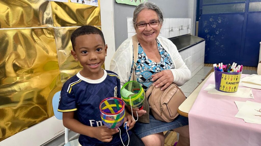A imagem mostra um menino e uma senhora sorridentes, sentados lado a lado em um ambiente que parece ser uma sala de aula ou um espaço de atividades recreativas. * O menino, que está à esquerda, veste uma camisa de futebol azul marinho com detalhes em amarelo e branco. Ele segura nas mãos dois brinquedos artesanais feitos de garrafas plásticas cortadas, decorados com fitas coloridas e barbantes. * A senhora, à direita, usa óculos, uma blusa estampada em tons de azul e branco, e um cardigã branco. Ela tem uma bolsa bege a tiracolo e também segura um dos brinquedos artesanais. * No canto direito, sobre uma mesa coberta por uma toalha rosa, há um recipiente roxo com o rótulo "CANETINHAS", contendo vários marcadores coloridos. O fundo apresenta uma parede parcialmente coberta por um papel dourado brilhante e um bebedouro ao fundo. A atmosfera é de alegria e interação.