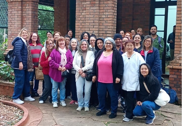 A imagem mostra um grupo de cerca de 25 pessoas, predominantemente mulheres, de diferentes idades, posando ao ar livre em frente a um edifício de tijolos vermelhos. Elas estão organizadas em várias fileiras, algumas em pé e outras um pouco adiantadas, formando uma composição coletiva. A maioria veste roupas casuais e apropriadas para clima fresco, como casacos, suéteres e cachecóis. As cores das roupas variam bastante, com destaque para uma blusa rosa no centro do grupo. O ambiente parece ser uma área externa pavimentada com pedras, possivelmente um pátio, e o céu está nublado, sugerindo iluminação natural difusa. A expressão das pessoas é majoritariamente sorridente ou neutra, e todas olham para a câmera. Ao fundo, além da parede de tijolos, é possível ver detalhes arquitetônicos como janelas e uma estrutura verde, possivelmente uma treliça. No canto direito inferior, há uma bolsa azul no chão.